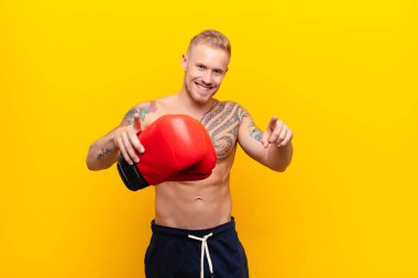 Genç sarışın, güçlü, mutlu ve kendinden emin bir adam. Kamerayı iki eliyle gösteriyor ve gülüyor. Seni boks eldivenleriyle seçiyor.