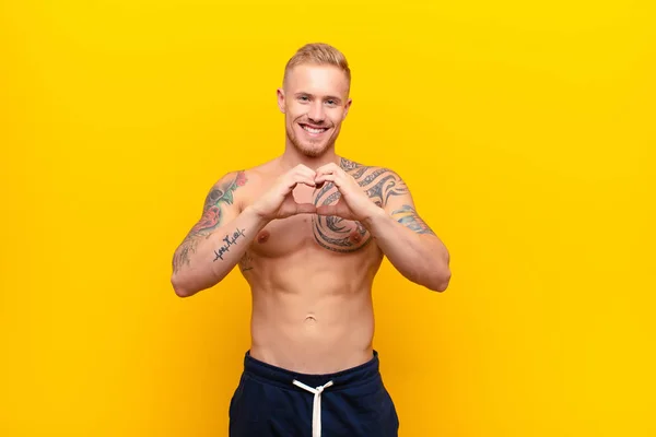 young blonde strong man smiling and feeling happy, cute, romantic and in love, making heart shape with both hands against yellow wall