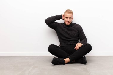 Sarışın genç adam kendini cahil ve kafası karışık hissediyor, bir çözüm düşünüyor, eli kalçasında, diğeri kafasında, dikiz aynası çimento zeminde oturuyor.