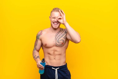 Genç sarışın güçlü adam mutlu bir şekilde gülümsüyor, şaka yapıyor ve gözetleme deliğinden bakıyor, enerji içeceğiyle sırları gözetliyor.