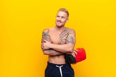 Genç sarışın güçlü bir adam mutlu, gururlu ve umutlu, merak eden ya da düşünen boks eldivenleriyle çapraz kollarıyla boşluğu arayan.