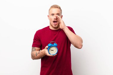 Genç sarışın adam mutlu, heyecanlı ve pozitif hissediyor. Elleri ağızda, beyaz duvarın karşısında çalar saati tutarak bağırıyor.