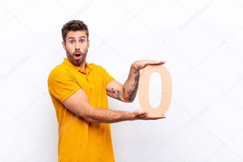 young handsome man surprised, shocked, amazed, holding a number 0.