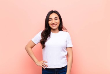 Genç Latin güzel kadın mutlu bir şekilde gülümsüyor. Kalça ve kendine güvenen, pozitif, gururlu ve düz duvara karşı dost canlısı bir tavırla.