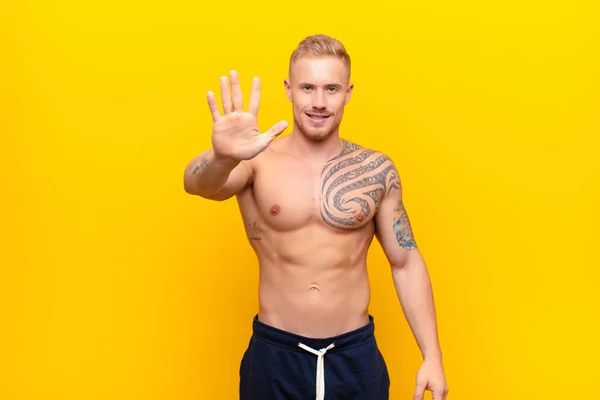 young blonde strong man smiling and looking friendly, showing number five or fifth with hand forward, counting down against yellow wall