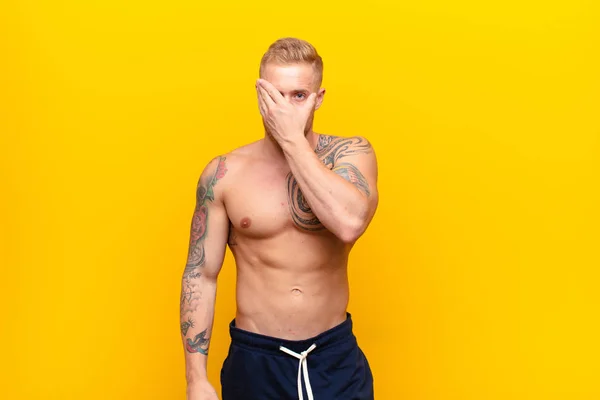 young strong blonde man feeling scared or embarrassed, peeking or spying with eyes half-covered with hands against yellow wall