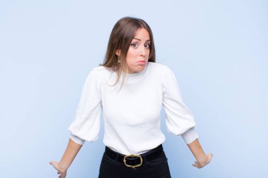young pretty hispanic woman feeling clueless and confused, having no idea, absolutely puzzled with a dumb or foolish look against blue wall