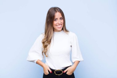 Genç, güzel, İspanyol bir kadın neşeli ve tesadüfen gülümsüyor. Mavi duvara karşı pozitif, mutlu, kendinden emin ve rahat bir ifadeyle.