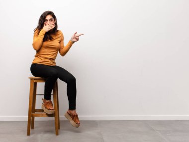 Genç ve güzel bir kadın mutlu, neşeli, şanslı ve şaşkın görünüyor. İki eli de bir odada otururken ağzını kapatıyor.