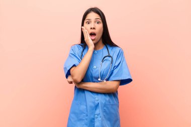 Genç Latin hemşire şok ve inançsızlıkla ağzı açık, elleri yanak ve kolları çaprazlanmış, sersemlemiş ve pembe duvara karşı hayrete düşmüş.