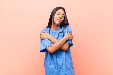 Genç Latin hemşire aşık oluyor, gülümsüyor, sarılıyor ve kendine sarılıyor, bekar kalıyor, bencil ve benmerkezci davranıyor pembe duvara yaslanıyor.