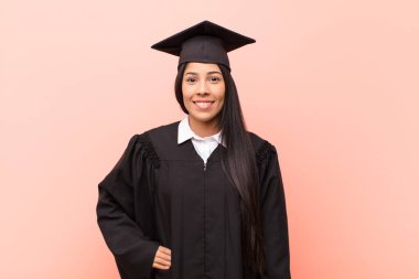 Genç Latin kız öğrenci mutlu bir şekilde gülümsüyor, elleri kalçasında ve kendinden emin, olumlu, gururlu ve arkadaş canlısı.