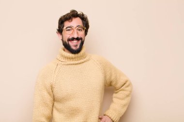 Havalı genç adam mutlu bir şekilde gülümsüyor, eli kalçasında ve kendinden emin, pozitif, gururlu ve dost canlısı bir tavırla düz duvara karşı.