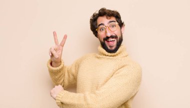 Genç, havalı bir adam gülümsüyor ve mutlu görünüyor, kaygısız ve pozitif, bir eliyle düz duvara karşı zafer ya da barış işareti yapıyor.