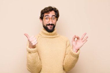 Havalı genç adam kendini mutlu, şaşkın, tatmin olmuş ve şaşırmış hissediyor, tamam gösteriyor ve jestler yapıyor, düz duvara gülümsüyor.