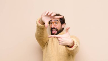 Havalı genç adam mutlu, arkadaş canlısı ve pozitif hissediyor, gülümsüyor ve elleri düz duvara dayalı bir portre veya fotoğraf çerçevesi yapıyor.