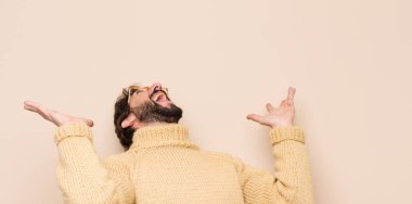 Genç havalı adam kendini mutlu, şaşkın, şanslı ve şaşırmış hissediyor, zaferi kutlarken iki elini de havaya kaldırmış düz duvara karşı.