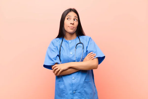 young latin nurse shrugging, feeling confused and uncertain, doubting with arms crossed and puzzled look against pink wall