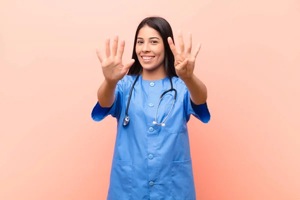若いラテン語の看護師は笑顔で友好的ですピンクの壁にカウントダウンしながら前方に手で9番目または9番目を示します — ストック写真