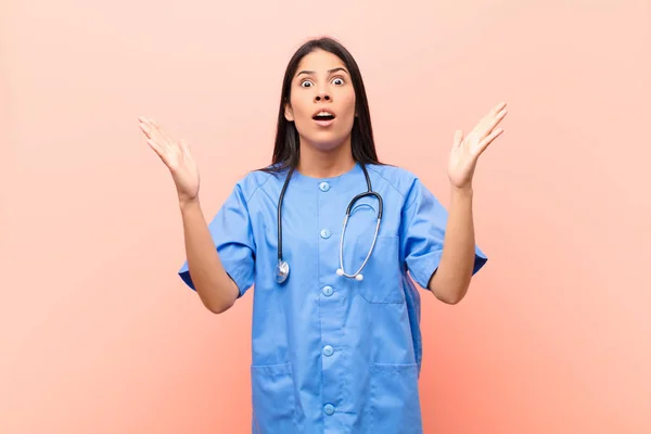 young latin nurse looking shocked and astonished, with jaw dropped in surprise when realizing something unbelievable against pink wall