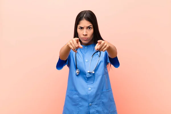 young latin nurse pointing forward at camera with both fingers and angry expression, telling you to do your duty against pink wall