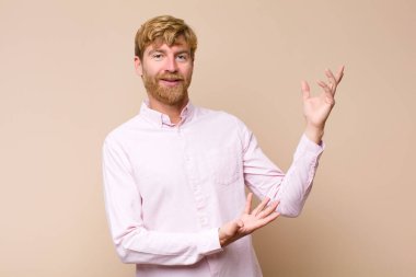 Genç sarışın adam gururla ve güvenle gülümsüyor, mutlu ve tatmin olmuş hissediyor ve düz duvara karşı fotokopi alanında bir konsept gösteriyor.