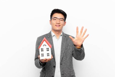 Genç Japon adam gülümsüyor ve arkadaş canlısı görünüyor, 5 ya da 5 numarayı gösteriyor, bir ev modeliyle geriye doğru sayıyor.