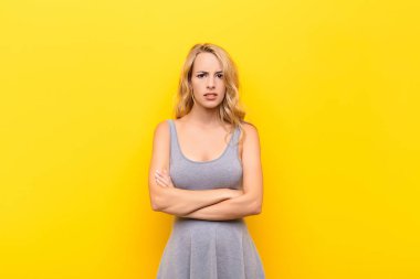 Genç sarışın kadın hayal kırıklığına uğramış, hayal kırıklığına uğramış, ciddi görünüyor, kızgın ve kızgın kollarını turuncu duvara dayamış.