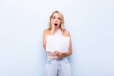 Genç sarışın kadın şok olmuş ve korkmuş hissediyor, ağzı açık ve elleri yanaklarında bir kağıt tutarak korkmuş görünüyor.