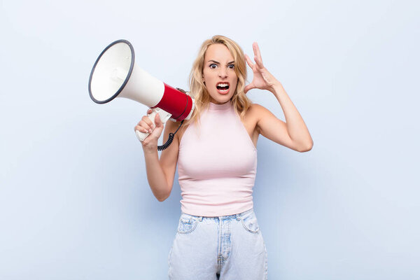 young blonde woman screaming with hands up in the air, feeling furious, frustrated, stressed and upset with a megaphone