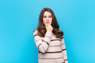 Genç ve güzel bir kadın ciddi, düşünceli ve güvensiz görünüyor, bir kolu çapraz ve çene üzerinde, mavi duvara karşı seçenekleri tartıyor