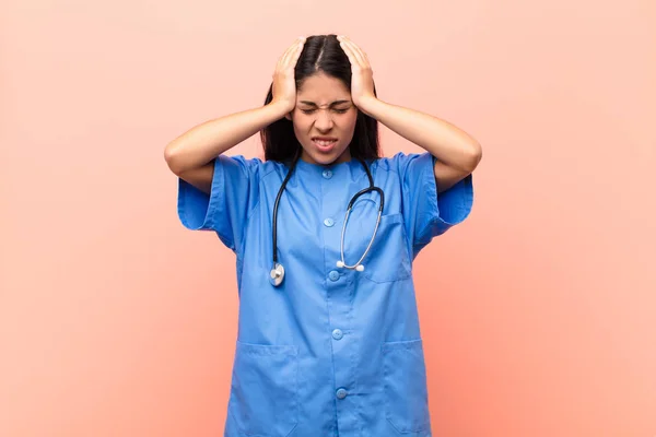 young latin nurse feeling stressed and frustrated, raising hands to head, feeling tired, unhappy and with migraine against pink wall