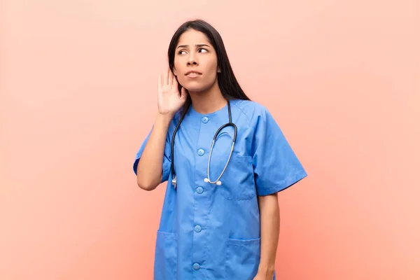 young latin nurse looking serious and curious, listening, trying to hear a secret conversation or gossip, eavesdropping against pink wall