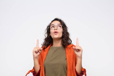 Genç ve güzel bir kadın şaşkınlık ve ağzı açık bir şekilde beyaz duvara karşı şaşkın ve şaşkın bir bakışla yukarıya bakıyor.