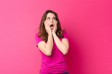 Genç, güzel bir kadın mutlu, heyecanlı ve şaşırmış hissediyor, yüzünü pembe duvara dayamış bir şekilde yan tarafa bakıyor.