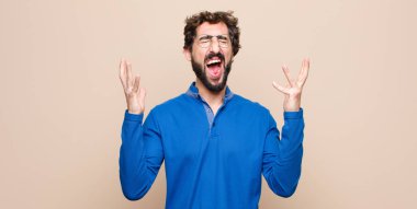 young handsome man furiously screaming, feeling stressed and annoyed with hands up in the air saying why me against flat wall
