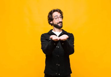 Genç çılgın yakışıklı adam mutlu bir şekilde gülümsüyor, arkadaş canlısı, kendinden emin, pozitif bakışlı, turuncu duvara karşı bir nesne ya da konsept sunuyor ve gösteriyor.