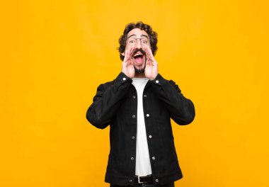 Genç çılgın yakışıklı adam mutlu, heyecanlı ve pozitif hissediyor. Elleri ağızda, turuncu duvara karşı bağırıyor.