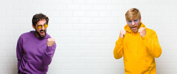 young cool men shouting aggressively with annoyed, frustrated, angry look and tight fists, feeling furious against white tiles wall