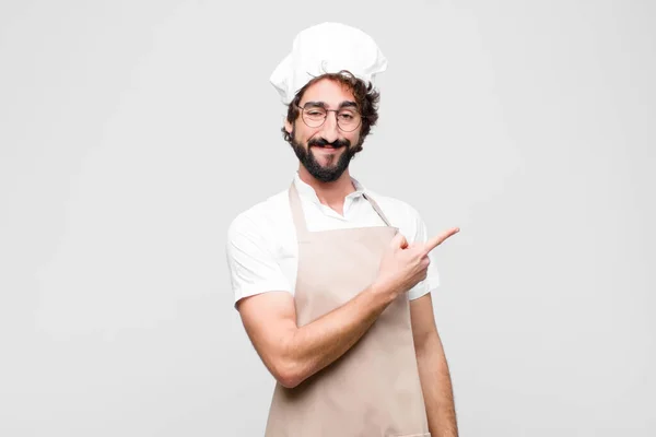 young crazy chef smiling cheerfully, feeling happy and pointing to the side and upwards, showing object in copy space against white wall