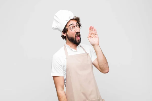 young crazy chef yelling loudly and angrily to copy space on the side, with hand next to mouth against white wall