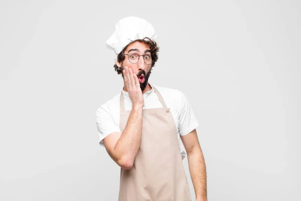 young crazy chef feeling shocked and astonished holding face to hand in disbelief with mouth wide open against white wall