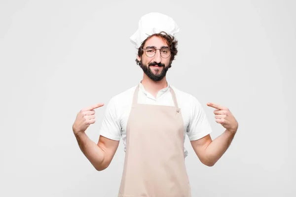 young crazy chef looking proud, positive and casual pointing to chest with both hands against white wall