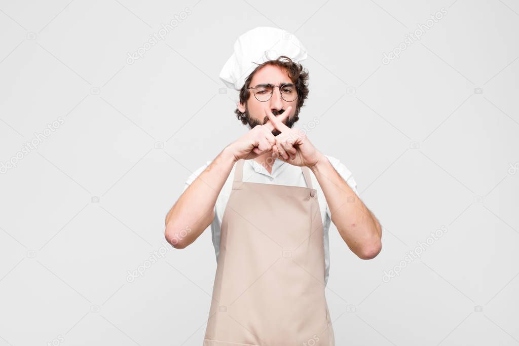 young crazy chef looking serious and displeased with both fingers crossed up front in rejection, asking for silence against white wall