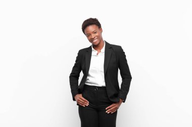 Genç, güzel, siyah bir kadın neşeli ve tesadüfen gülümsüyor. Beyaz duvara karşı pozitif, mutlu, kendinden emin ve rahat bir ifadeyle.