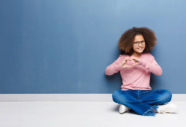 アフリカ系アメリカ人の少女は笑顔で幸せを感じ 可愛くてロマンチックで愛を感じ 両手で床に座ってハートの形を作っています — ストック写真