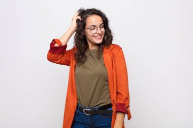 Genç, güzel bir kadın neşeli ve tesadüfen gülümsüyor, beyaz duvara karşı pozitif, mutlu ve kendinden emin bir bakışla göğüs göğüse tutuşuyor.