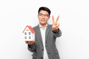 Genç Japon adam gülümsüyor ve mutlu görünüyor, kaygısız ve pozitif, bir ev modeliyle zafer veya barış işareti yapıyor.