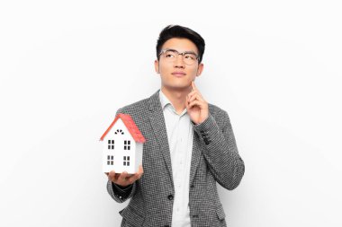 Genç Japon adam mutlu bir şekilde gülümsüyor ve hayal kuruyor ya da şüphe ediyor, bir ev modeliyle yan yana bakıyor.