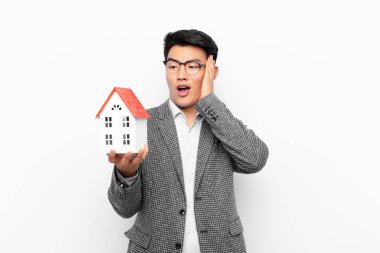 Genç Japon bir adam mutlu, heyecanlı ve şaşırmış hissediyor, iki eli yüzü üzerinde bir ev modeliyle yan tarafa bakıyor.